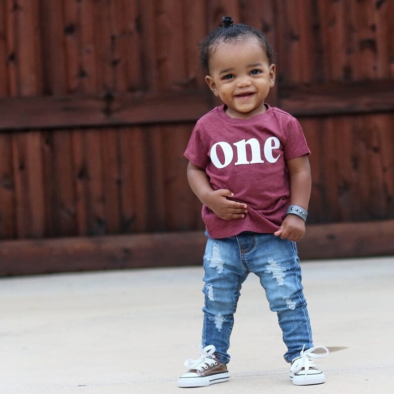 May include: A young child wearing a maroon t-shirt with the word "one" printed in white. The child is wearing ripped denim jeans and brown and white sneakers. The child is standing in front of a wooden fence.
