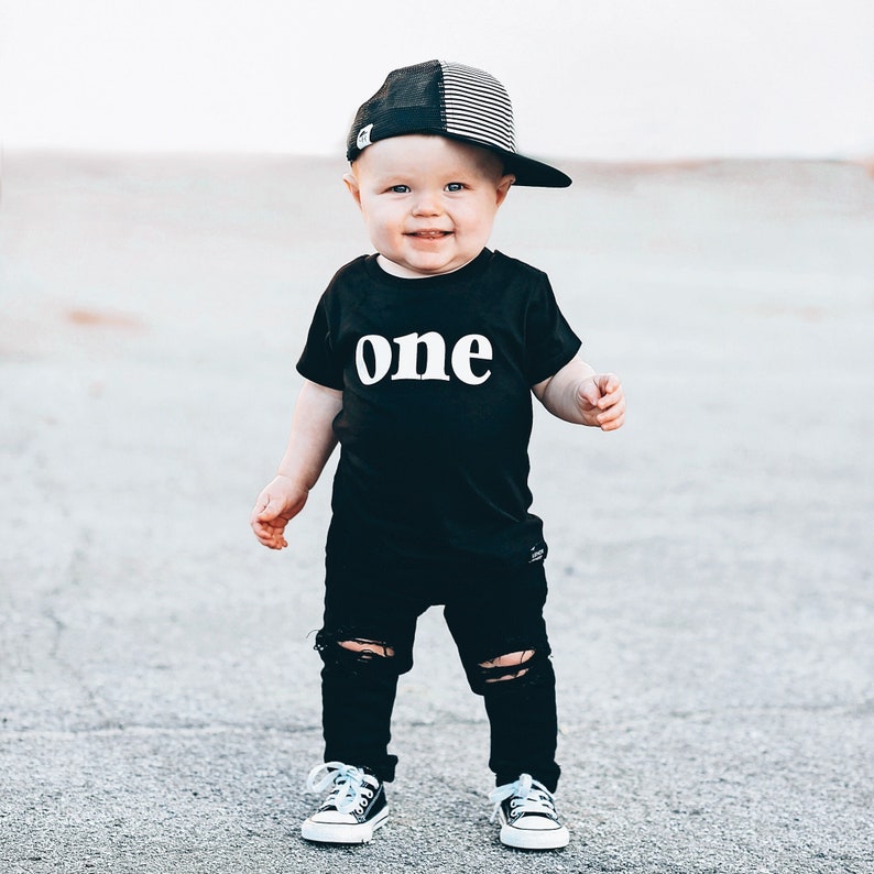 May include: A toddler wearing a black t-shirt with the word "one" in white, black ripped jeans, black and white trucker hat, and blue and white sneakers.