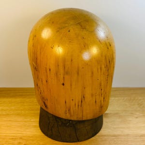 May include: A smooth, light-colored wooden hat block shaped like a human head, featuring natural wood grain patterns and dark streaks. The head form is mounted on a dark, cylindrical wooden base with a visible vertical crack in the wood grain.