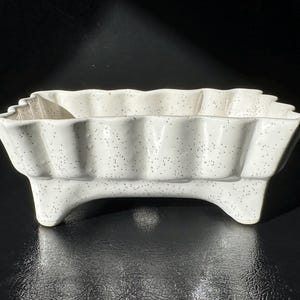 May include: A white ceramic planter with a scalloped rim and speckled finish. The rectangular planter sits on three curved feet. The planter is suitable for indoor or outdoor use.