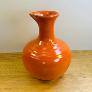 May include: A bright orange ceramic vase with a smooth, glossy finish. The vase features a rounded, bulbous body that tapers into a slender neck, ending in a flared, circular rim. The surface reflects light, highlighting the vibrant orange color.