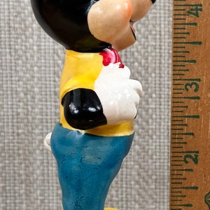 May include: A ceramic figurine of Mickey Mouse wearing a yellow shirt, blue jeans, and yellow shoes. He is standing on a blue base.