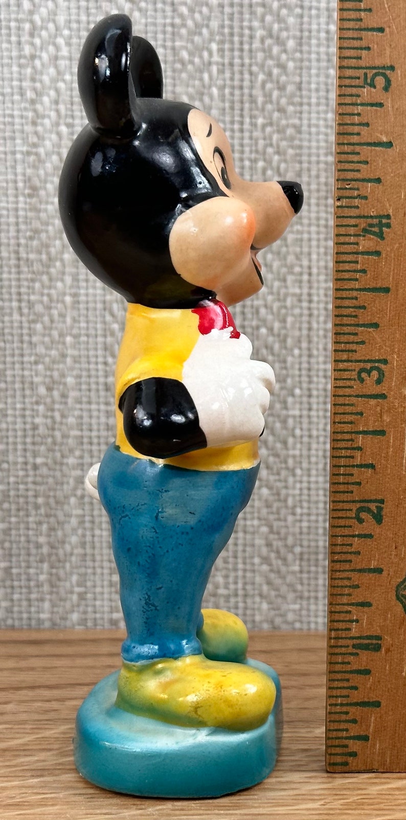 May include: A ceramic figurine of Mickey Mouse wearing a yellow shirt, blue jeans, and yellow shoes. He is standing on a blue base.