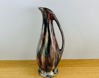May include: A small, ceramic vase with a brown, black, and white swirled glaze. The vase has a rounded body and a narrow neck with a spout. It has a single handle.