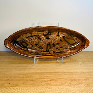 May include: Oval-shaped ceramic serving dish with a brown and gold glaze. The dish features a textured, organic design in the center. The rim has a darker brown color. The dish is displayed on a clear stand.