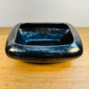 May include: A dark, square-shaped bowl with rounded corners. The bowl has a metallic, iridescent finish with shades of blue and green. The bowl is sitting on a wooden surface.