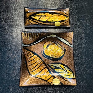May include: Two decorative glass trays with leaf designs. The larger tray is square, and the smaller one is rectangular. Both have a brown and gold color scheme with black accents. The leaf designs are in shades of gold and yellow.