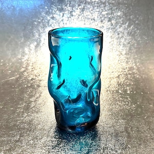 May include: A turquoise-colored, hand-blown glass tumbler with a unique, textured design. The glass has an organic, wavy pattern, and the open top is smooth. The tumbler is approximately 4 inches tall and is suitable for beverages.