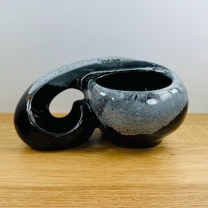 May include: A black and white ceramic planter with a unique, abstract design. The planter has a large, open bowl and a curved handle.