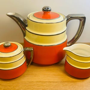May include: A vintage tea set with a teapot, sugar bowl, and creamer. The set features a color scheme of orange, yellow, and silver. The teapot has a lid with a silver knob and a silver handle. The sugar bowl has a lid and handles. The creamer has a handle.