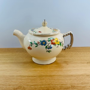 May include: A cream-colored teapot with a silver handle and lid. The teapot is decorated with a floral pattern in shades of blue, yellow, red, and green.