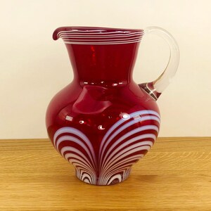 May include: A red glass pitcher with white swirling lines. The pitcher has a clear glass handle.