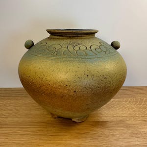 May include: A round ceramic vessel with a wide body, a short neck, and a dark rim. The surface features a speckled mustard yellow and green gradient finish with an embossed swirling pattern near the top and two small spherical side handles.