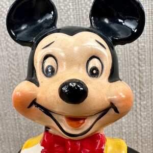May include: A ceramic figurine of Mickey Mouse wearing a yellow shirt, red bow tie, and white gloves. He has a big smile and large black ears.