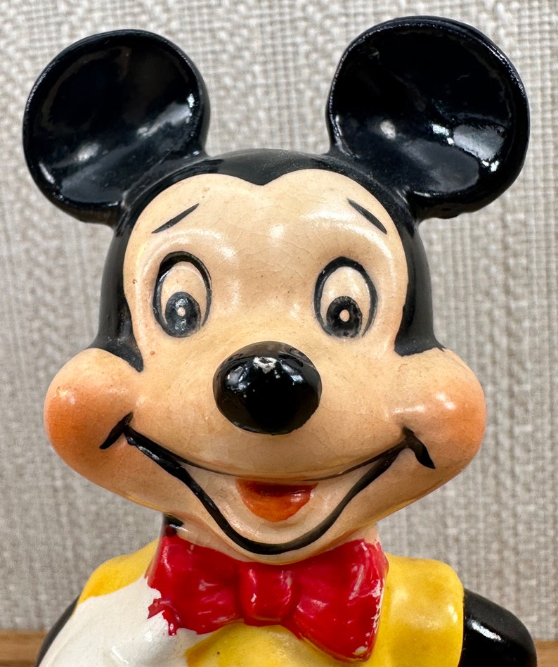 May include: A ceramic figurine of Mickey Mouse wearing a yellow shirt, red bow tie, and white gloves. He has a big smile and large black ears.