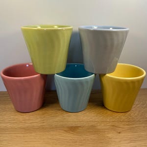 May include: A collection of five ceramic cups in various colors: yellow, pink, green, blue, and gray. Each cup has a unique, slightly swirled design and a tapered shape. The cups are arranged on a wooden surface.