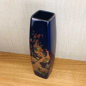 May include: A cobalt blue ceramic vase with a square opening and a rounded body. The vase is decorated with gold and red accents and features a design of two peacocks with their tails spread.