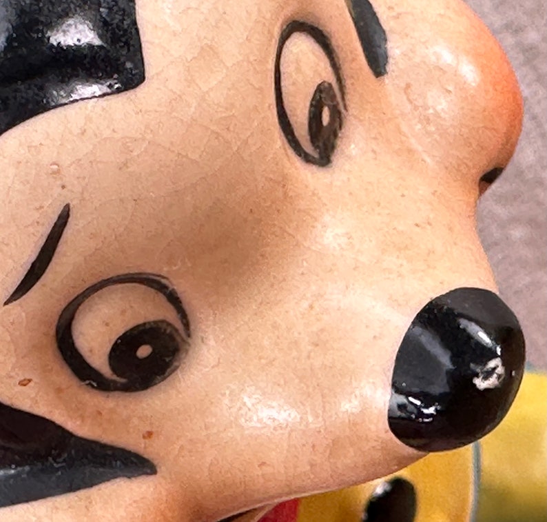 May include: Close-up of a ceramic figurine's face with black painted eyes, eyebrows, and a mustache. The figurine has a light brown skin tone and a black nose.