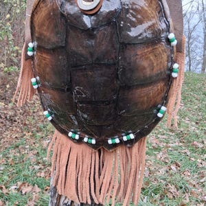 以下が含まれることがあります： 茶色の革のストラップとフリンジが付いた、カメの甲羅のバックパック。甲羅は濃い茶色で光沢があります。白と緑のビーズが縁に縫い付けられています。