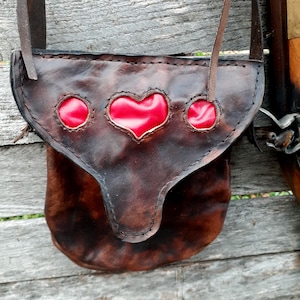 Puede incluir: Una bolsa de cuero marrón con cierre de solapa y tres recortes en forma de corazón rojo. La bolsa tiene una correa y se lleva sobre el hombro.