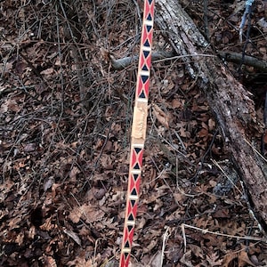 以下が含まれることがあります： 装飾的なデザインのロングボウ。弓は、その長さに沿って赤、黒、白の幾何学模様が特徴です。弓は木製で、革製のグリップが付いています。背景は、葉や枝のある自然な環境です。