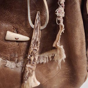 May include: A brown leather bag featuring a light-colored toggle closure and a carved pendant hanging from a thin cord. The pendant displays a detailed face with a beard, and the bag has a frayed, raw-edged pocket flap with visible stitching.