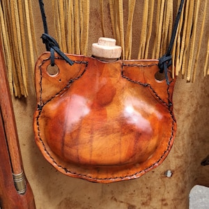 Puede incluir: Una cantimplora de cuero hecha a mano con un tapón de madera y un cordón negro para llevar. La cantimplora tiene forma redondeada y está cosida con hilo negro. El cuero es de color marrón rojizo cálido.