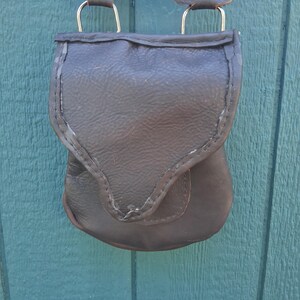 May include: A dark brown leather pouch with a pointed flap closure, featuring visible stitching along the edges. The pouch is attached to a green fabric strap by two silver-toned metal D-rings connected to leather tabs at the top of the bag.