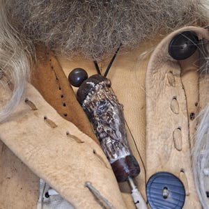 Könnte beinhalten: Eine handgefertigte Anhängerkette mit einem geschnitzten Geweihanhänger und einem dunkelbraunen Holzsockel. Die Kette hängt an einer schwarzen Kordel vor einer hellbraunen Lederjacke mit großen schwarzen Knöpfen.