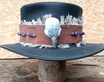 Puede incluir: Un sombrero fedora negro con ala ancha. Cuenta con una banda de cuero marrón adornada con cuentas azules y blancas y una calavera de pájaro plateada. El ala está adornada con tela deshilachada.