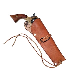 Könnte beinhalten: Ein Revolver im Vintage-Stil mit Holzgriff und Messingrahmen, der teilweise in einem hellbraunen Lederholster steckt. Das Holster verfügt über einen Schnallenriemen, einen einzelnen Metallniet und lange Lederbänder, die unten herausragen.