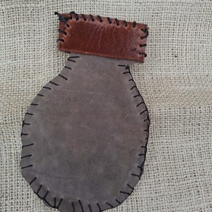 May include: A handmade leather pouch with a brown top and a gray body. The pouch is stitched together with black thread. The top is rectangular, and the body is a rounded shape. The pouch is displayed on a burlap surface.