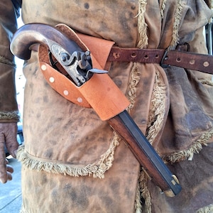 May include: A vintage pistol in a brown leather holster, attached to a brown leather belt. The pistol has a wooden handle and metal components. The person is wearing a brown jacket with fringe.