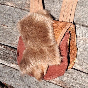 May include: A handmade leather pouch with a fur flap and tan straps. The pouch is a mix of reddish-brown and tan leather, with a textured surface. The fur is a light brown color. The straps are a light tan color with a stitched design.