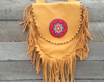 May include: A tan leather bag with fringe and a beaded design. The bag has a circular beaded design in the center with red, blue, white, and yellow beads.