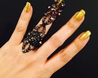 May include: A black, articulated finger ring with a filigree design and studded accents. The ring is worn on a hand with gold glitter nail polish. The background is black.