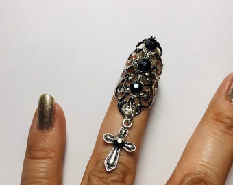 May include: Silver-toned finger ring with a decorative filigree design and black gemstone accents. A dangling cross pendant hangs from the ring. The ring is worn on a finger with gold nail polish.