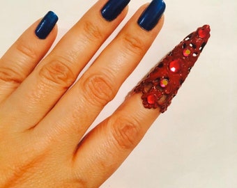 May include: A hand with dark blue nail polish and a decorative, reddish-brown finger cover adorned with red and clear embellishments. The finger cover has a floral design and is worn on the index finger.