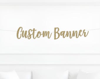 May include: A gold glitter "Custom Banner" strung across a white wall. The banner is in a cursive font and hangs above a white sofa with pillows. The wall has decorative molding.