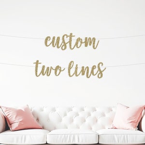 May include: Gold glitter banner with the text "custom two lines" hanging on a white wall above a white sofa with pink pillows and pink and white balloons scattered on the floor.