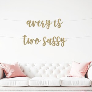 May include: A gold glitter banner with the text "avery is two sassy" hanging above a white sofa with pink pillows. Pink and white balloons are scattered on the floor in front of the sofa.