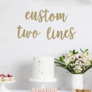 May include: A white table with a white tablecloth, a white cake with gold flecks, a white cake stand, cupcakes with red berries, a bouquet of white flowers in a white vase, and a gold glitter banner that says "custom two lines".