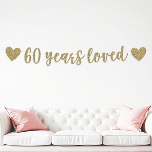 May include: Gold glitter banner with the text "60 years loved" and two gold glitter hearts on either side. The banner is hanging above a white sofa with pink pillows. There are pink and white balloons on the floor in front of the sofa.