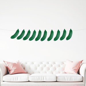 May include: A white tufted sofa with pink pillows and scattered balloons. A string of green cucumber-shaped decorations hangs above the sofa. The balloons are a mix of white and pink.