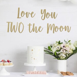 May include: A party scene with white balloons, a white cake, and a banner that reads "Love You TWO the Moon" in gold lettering. The table is set with desserts and flowers, creating a celebratory atmosphere.