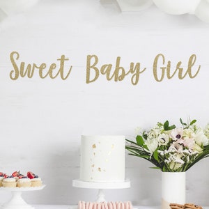 May include: A white table with a white tablecloth, a white cake with gold flecks, cupcakes with strawberries, a bouquet of white flowers in a white vase, and a banner that reads 'Sweet Baby Girl' in gold glitter letters.