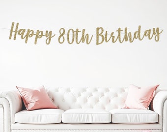 May include: Gold glitter banner with the text "Happy 80th Birthday" hanging above a white sofa with pink pillows and pink and white balloons scattered on the floor.