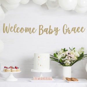 May include: A white table with a white tablecloth, a white cake, cupcakes with berries, and a bouquet of white flowers. A gold glitter banner reads "Welcome Baby Grace".
