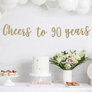 May include: A party scene with a white cake, cupcakes, and flowers on a table. A banner reading "Cheers to 90 years" hangs above. White balloons frame the scene, creating a festive atmosphere.