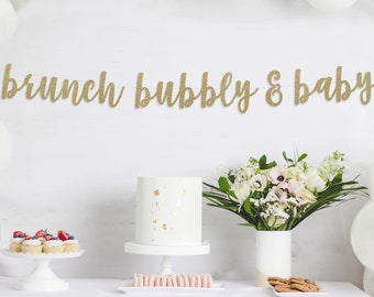 May include: A festive brunch and baby shower setup with white balloons, a white cake, and a banner that reads "brunch bubbly & baby." The table is set with desserts and flowers.